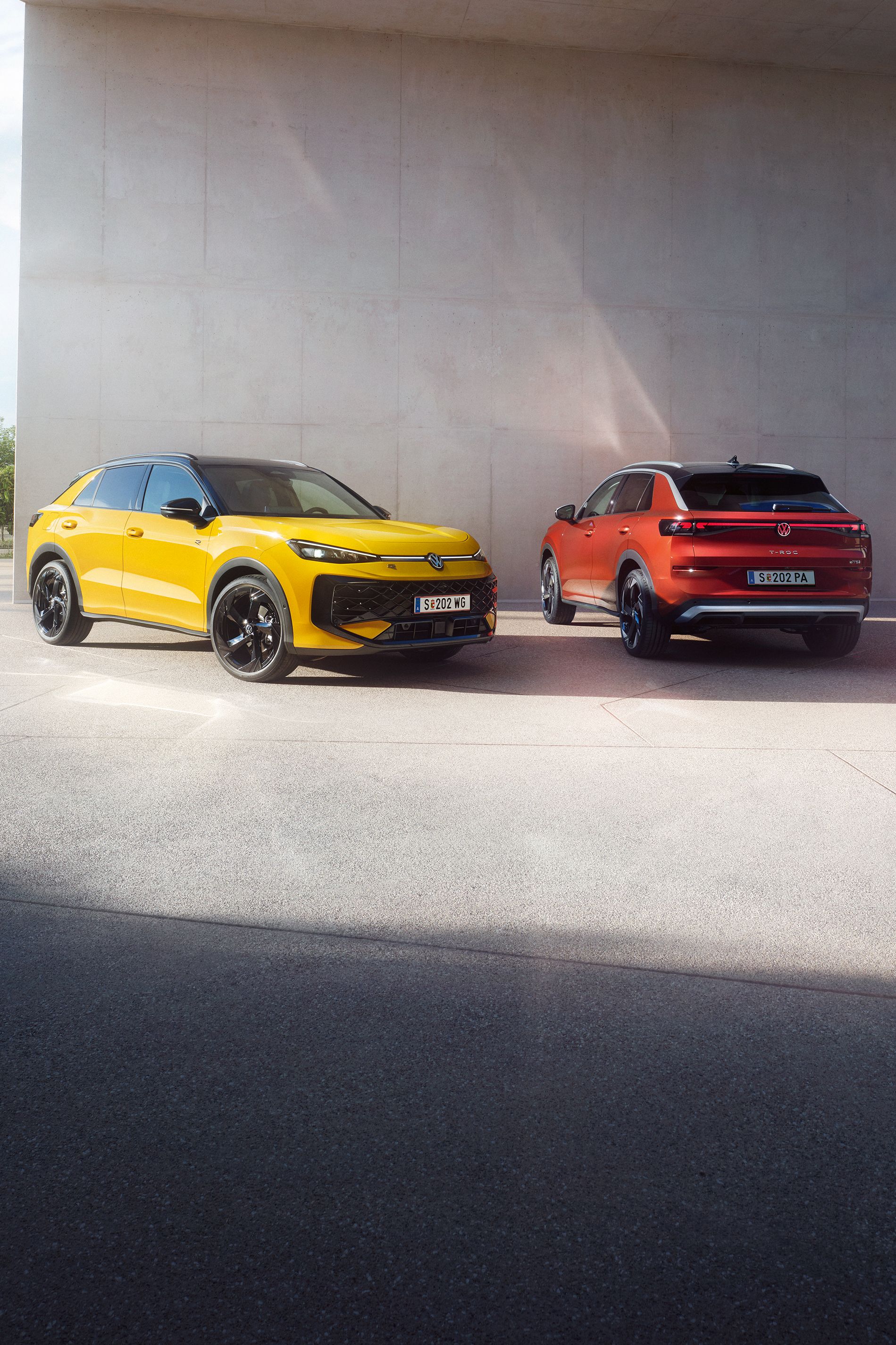 Ein gelber VW T-Roc R-Line und ein roter VW T-Roc Style stehen nebeneinander vor einem modernen Gebäude.