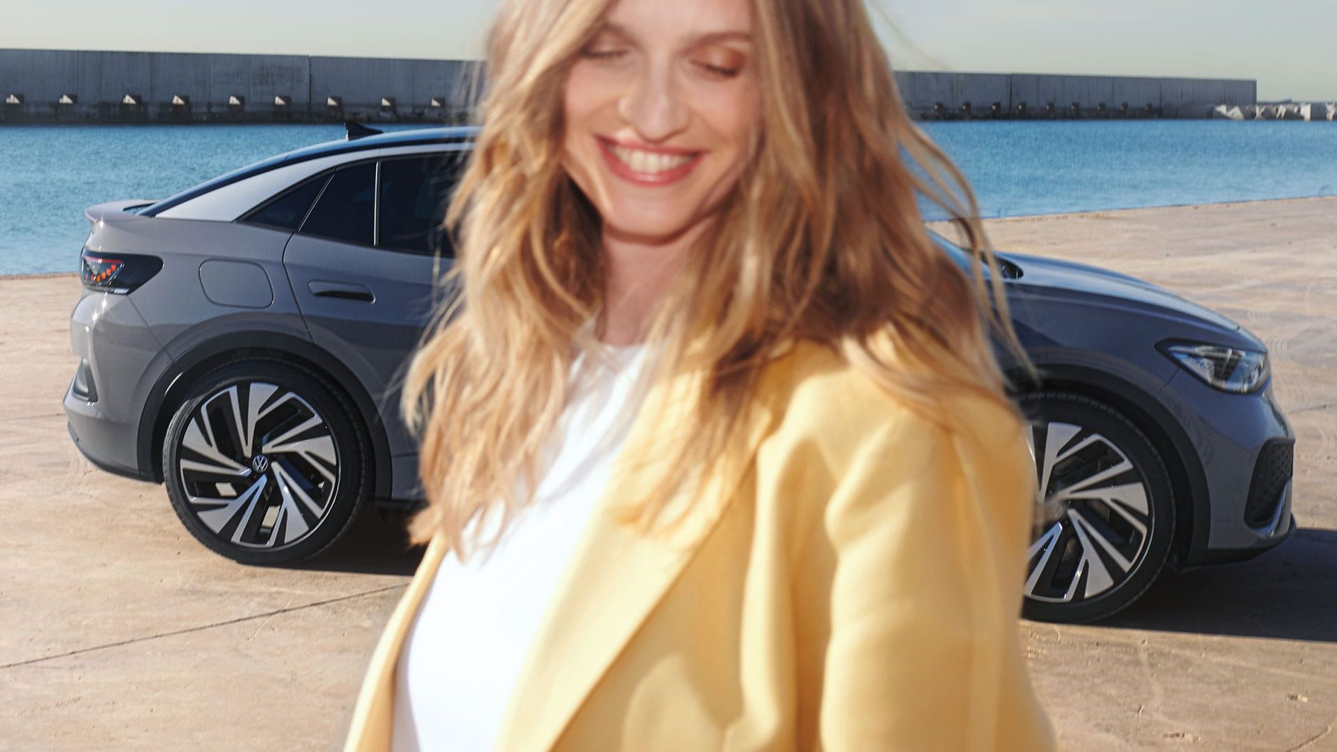 Seitenansicht eines grauen VW ID.5 an einem Hafenkai, im Vordergrund eine lächelnde, hell gekleidete Frau