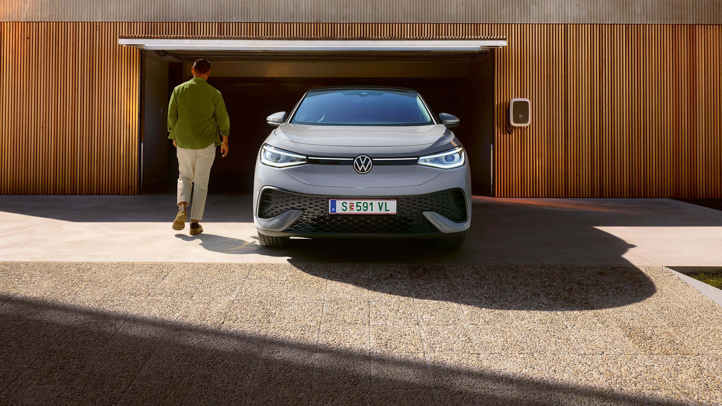 Grauer VW ID.5 von vorne vor einer Garage mit Wallbox, ein Mann geht am Auto vorbei