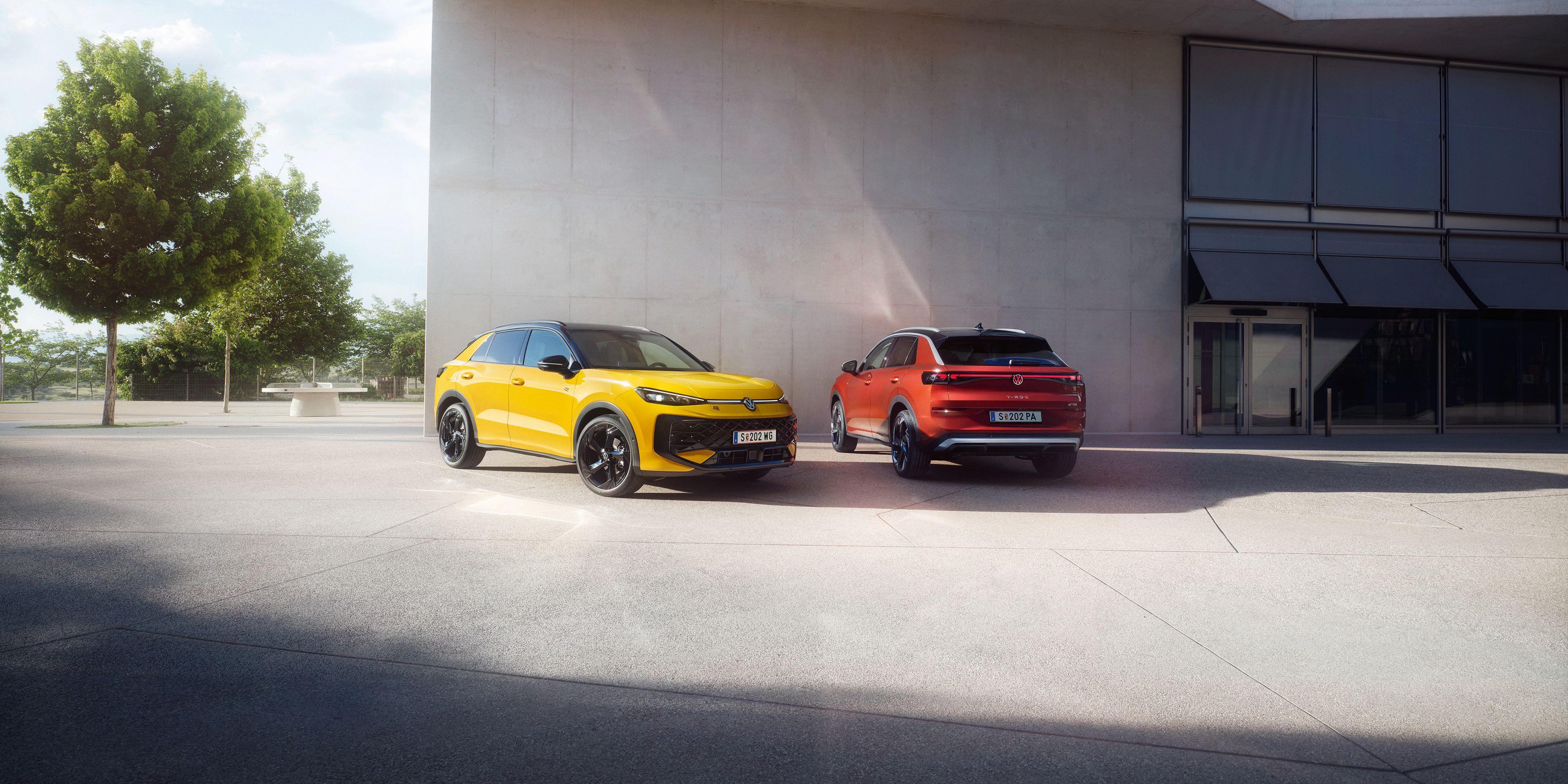 Ein gelber VW T-Roc R-Line und ein roter VW T-Roc Style stehen nebeneinander vor einem modernen Gebäude.